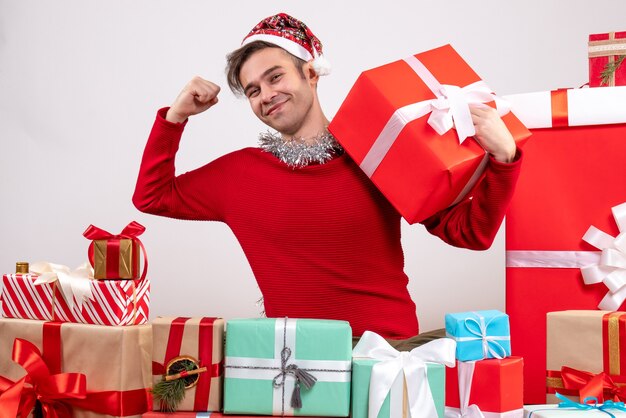 Вид спереди молодой человек показывает мышцы, сидя вокруг рождественских подарков