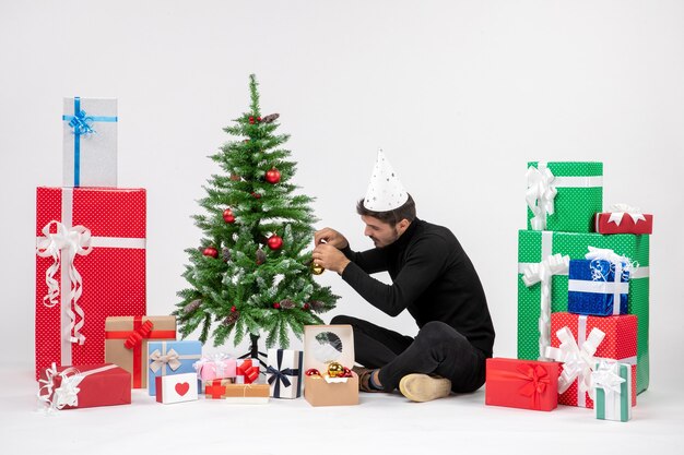 Front view of young man sitting around holiday presents decorating little tree on the white wall