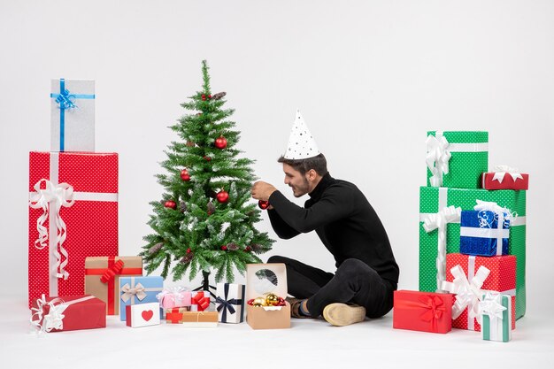 Front view of young man sitting around holiday presents decorating little tree on white wall