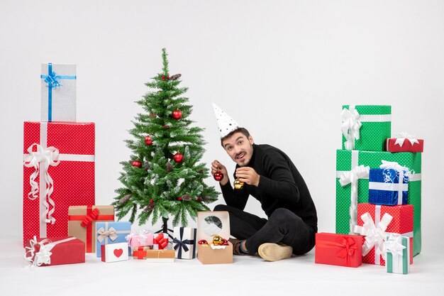 Front view of young man sitting around holiday presents decorating little tree on white wall