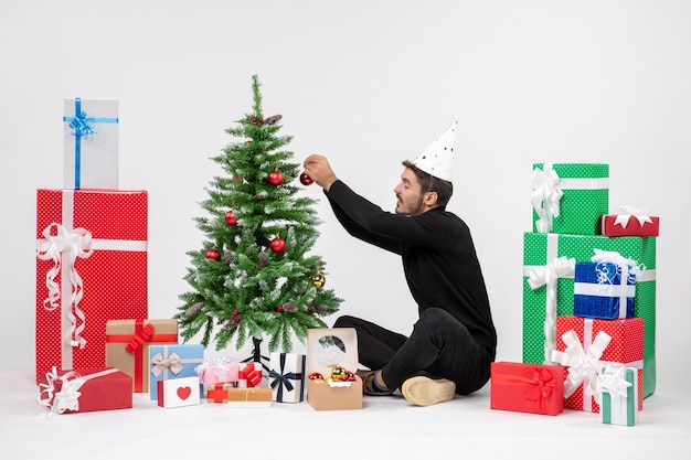 Front view of young man sitting around holiday presents decorating little tree on white wall