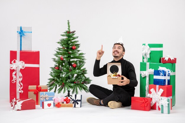 Front view of young man sitting around holiday presents holding package with toys on white wall