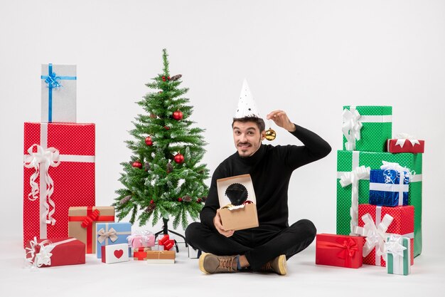 Front view of young man sitting around holiday presents holding toys on white wall