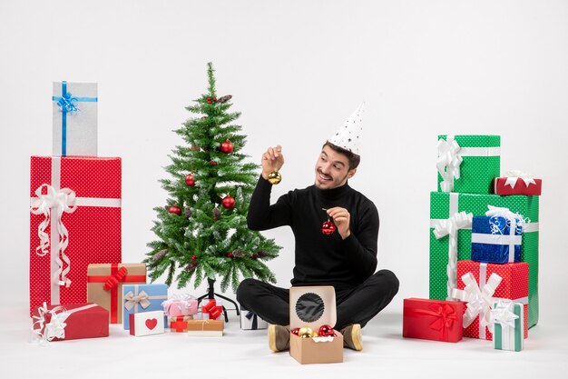 Вид спереди молодого человека, сидящего вокруг праздничных подарков, держащего елочные игрушки на белой стене