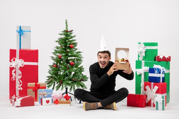 Front view of young man sitting around holiday presents holding tree toys on white wall