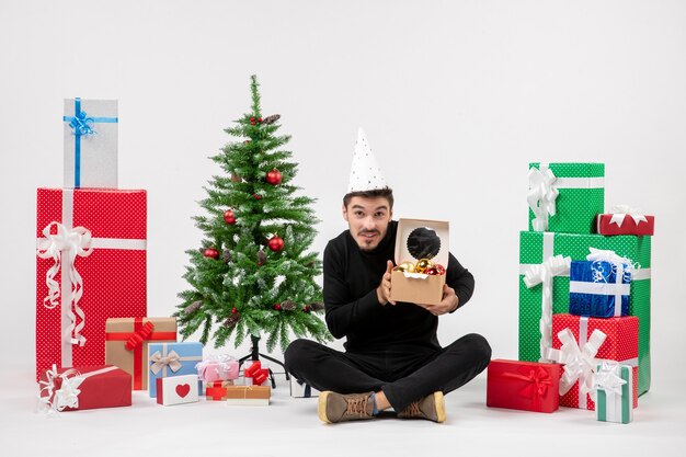 Front view of young man sitting around holiday presents holding tree toys on white wall