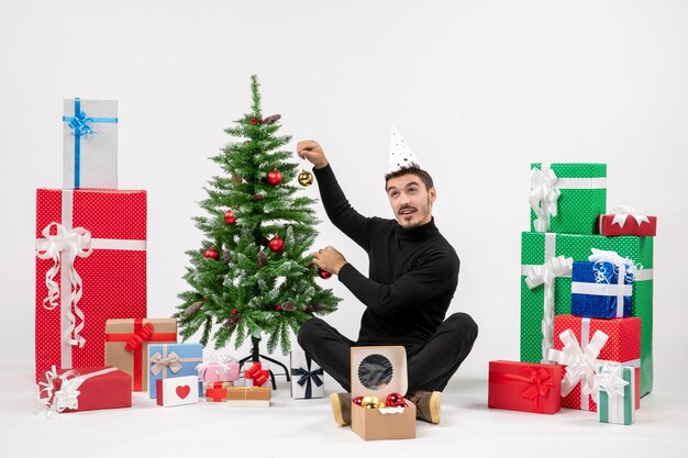 Front view of young man sitting around holiday presents holding tree toys on white wall