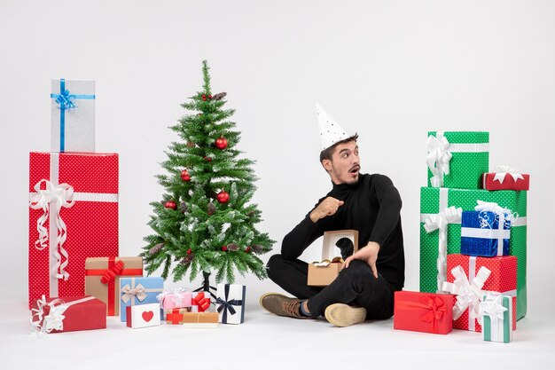 Front view of young man sitting around holiday presents on a white wall