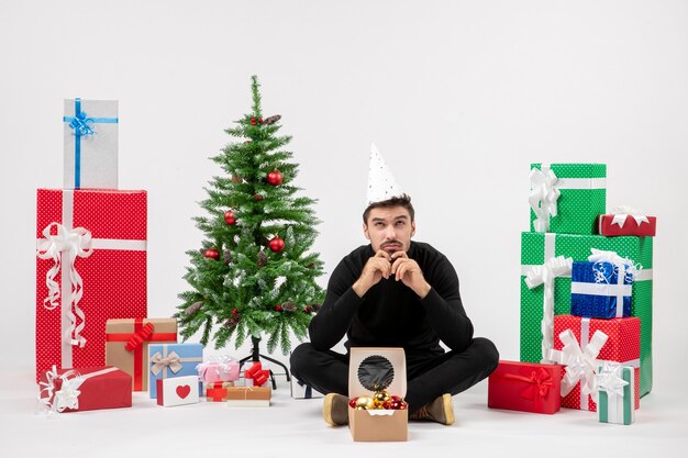 Front view of young man sitting around holiday presents on a white wall