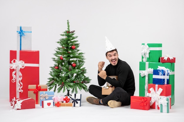 Front view of young man sitting around holiday presents on a white wall