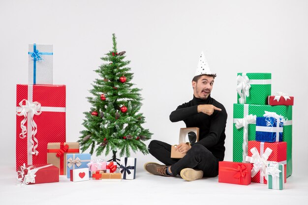 Front view of young man sitting around holiday presents on the white wall