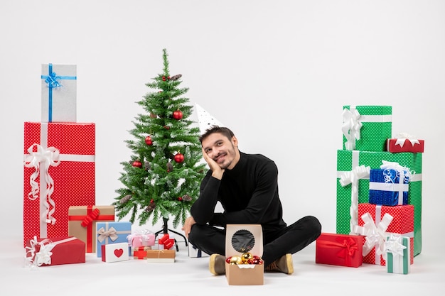 Front view of young man sitting around holiday presents on white wall