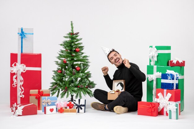 Front view of young man sitting around holiday presents on white wall