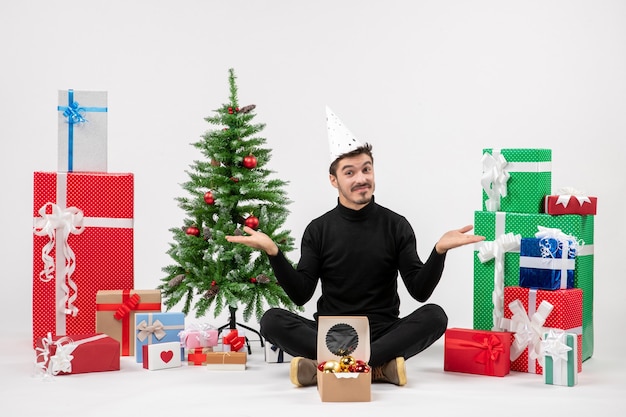 Вид спереди молодого человека, сидящего вокруг праздничных подарков на белой стене