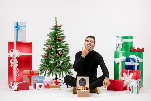 Free photo front view of young man sitting around holiday presents and yawning on white wall