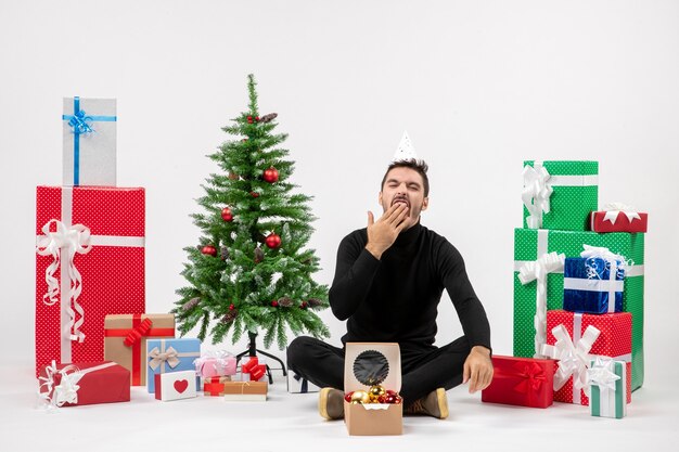Вид спереди молодого человека, сидящего вокруг праздничных подарков и зевающего на белой стене