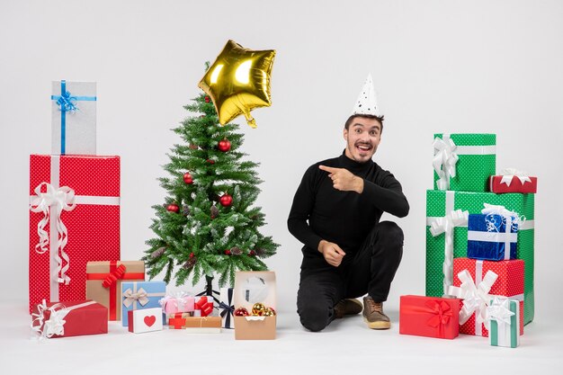 Front view of young man sitting around presents and gold star balloon on white wall