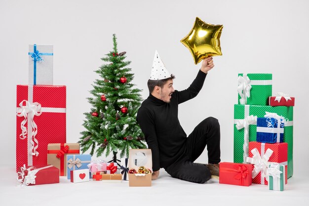 Front view of young man sitting around presents and holding gold star figure on white wall