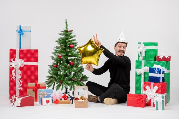 Front view of young man sitting around presents and holding gold star figure on white wall