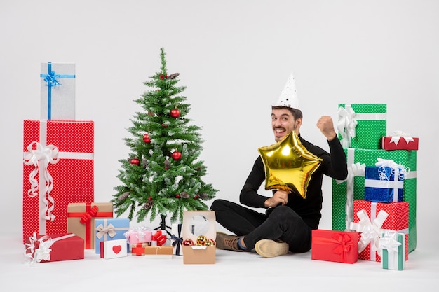 Front view of young man sitting around presents and holding gold star figure on white wall