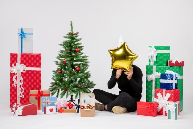 Front view of young man sitting around presents and holding gold star figure on white wall