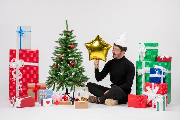 Front view of young man sitting around presents and holding gold star figure on white wall