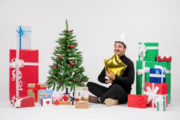 Front view of young man sitting around presents and holding gold star figure on white wall