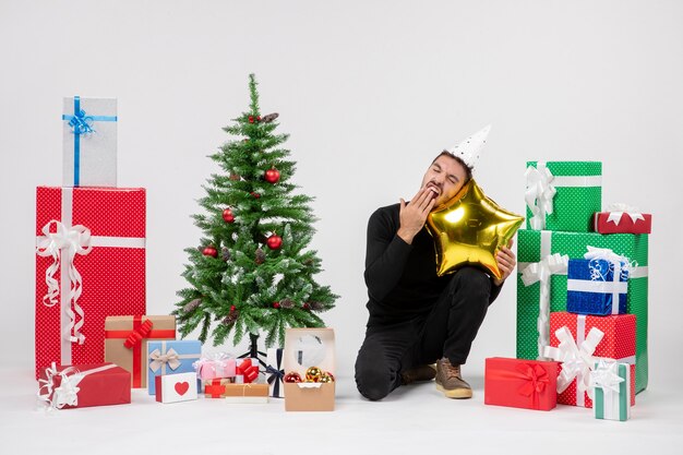 Front view of young man sitting around presents and holding gold star shaped pillow on white wall