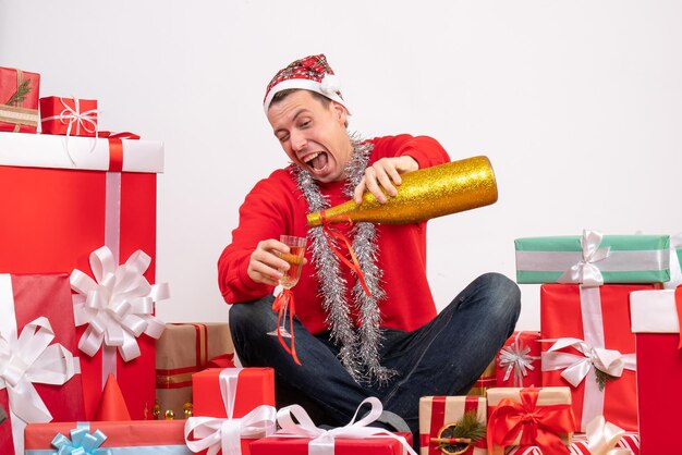Front view of young man sitting around presents pouring champagne on white wall