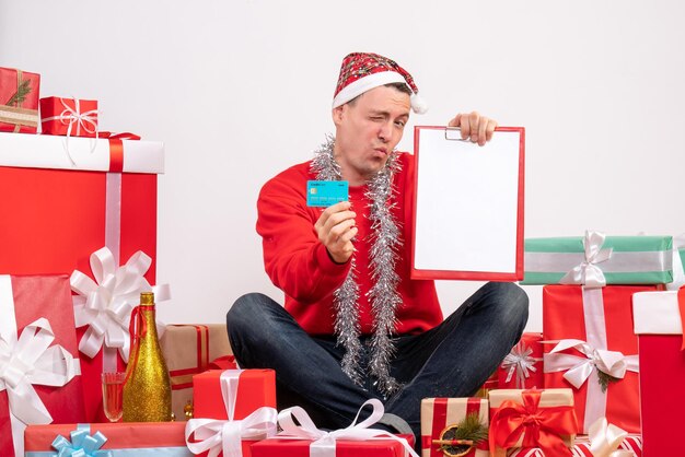 Front view of young man sitting around presents with bank card and note on white wall