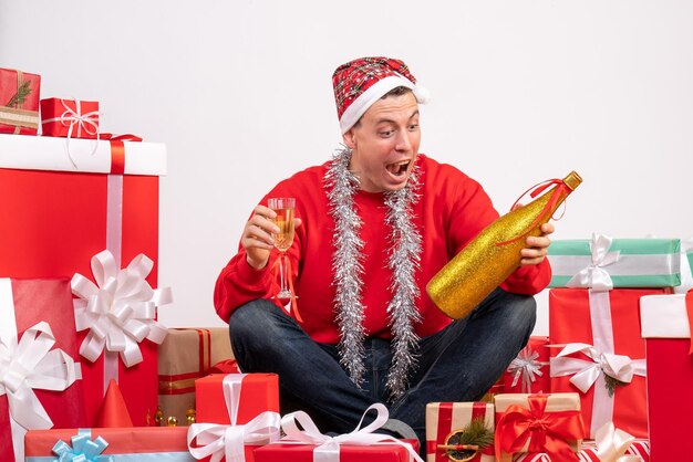Front view of young man sitting around presents with champagne on white wall