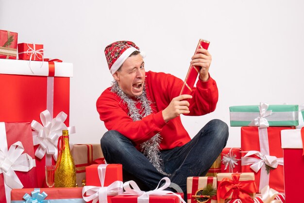 Front view of young man sitting around presents with firecracker on a white wall