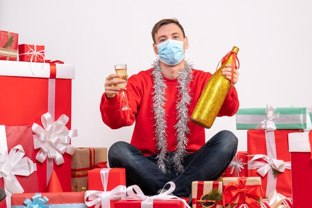 Вид спереди молодого человека, сидящего вокруг рождественских подарков, празднует с шампанским на белой стене