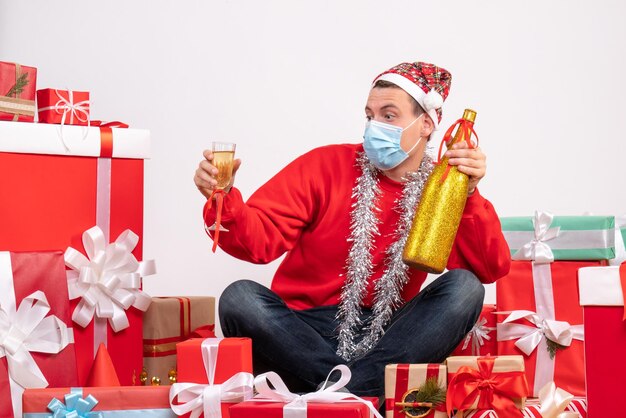 Front view of young man sitting around xmas presents celebrating with champagne on white wall