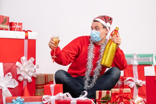 Вид спереди молодого человека, сидящего вокруг рождественских подарков, празднует с шампанским на белой стене