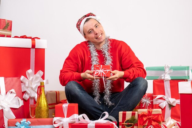 Front view of young man sitting around xmas presents on white wall