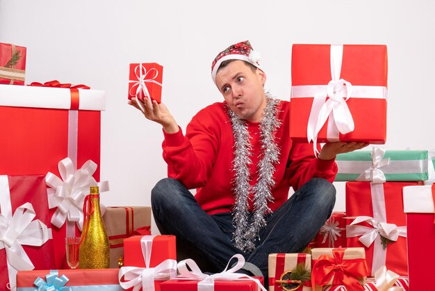 Front view of young man sitting around xmas presents on white wall