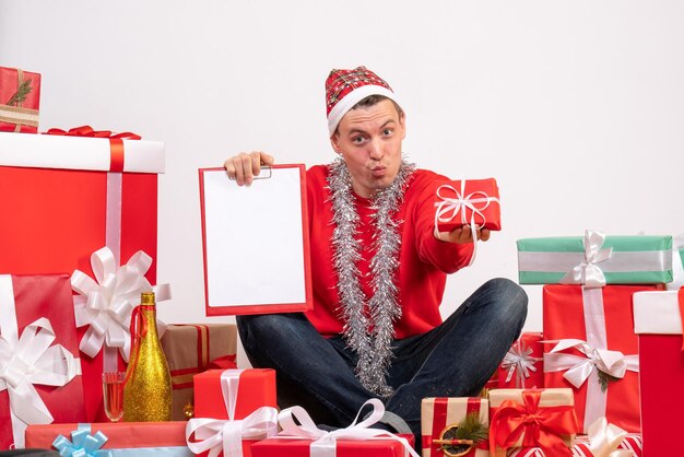 Front view of young man sitting around xmas presents with clipboard on white wall