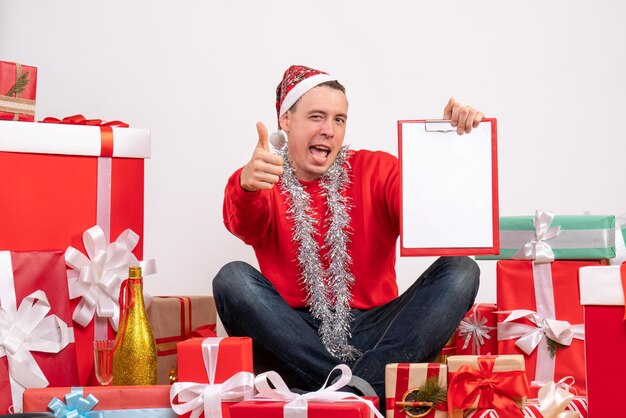 크리스마스 선물 주위에 앉아 있는 젊은 남자의 전면 뷰는 흰색 벽에 클립보드를 제공합니다.