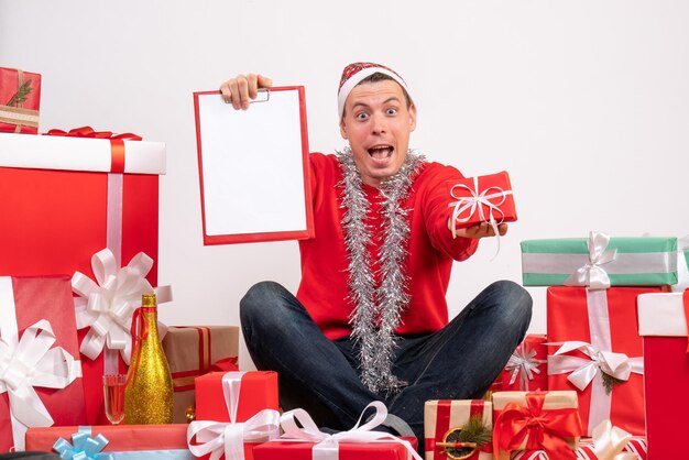 Вид спереди молодого человека, сидящего вокруг рождественских подарков с буфером обмена на белой стене