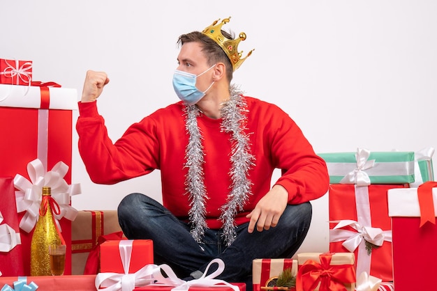 白い壁にクリスマスプレゼントの周りに座っている滅菌マスクの若い男の正面図