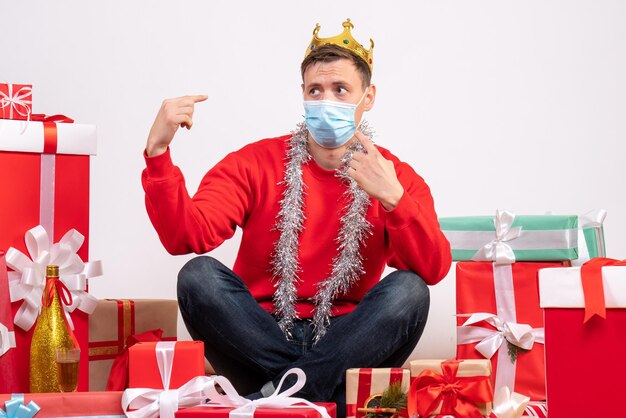 Вид спереди молодого человека в стерильной маске, сидящего вокруг рождественских подарков на белой стене