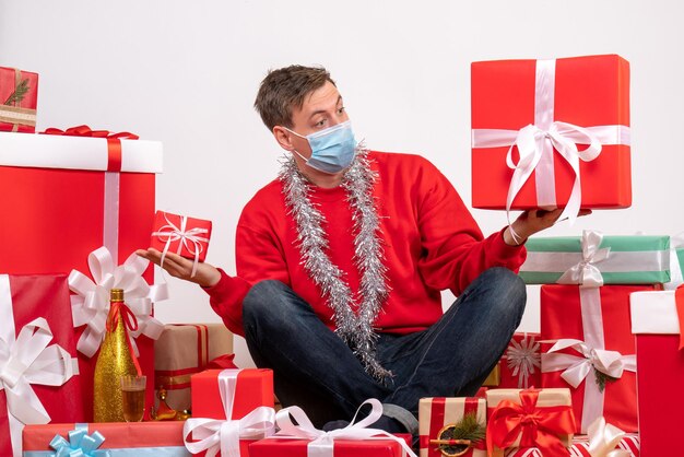 白い壁にクリスマスプレゼントの周りに座っている滅菌マスクの若い男の正面図