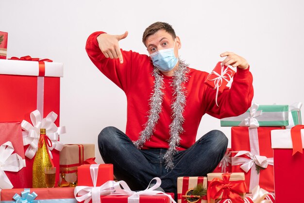 Front view of young man in sterile mask sitting around xmas presents on white wall