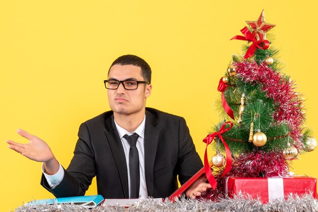 Front view young man in a suit sitting at the table opening his hands xmas tree and gifts