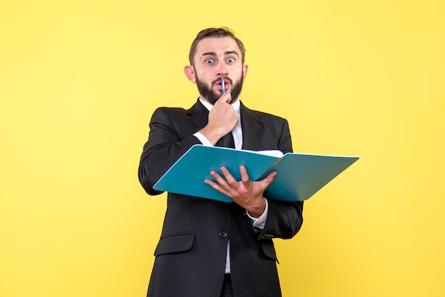Вид спереди удивленного молодого человека, бизнесмена, касающегося ручкой рта, держит синюю папку на желтом
