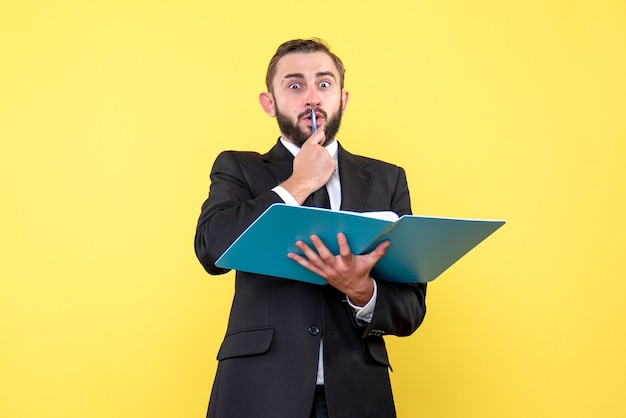 Front view of young man surprised businessman touching pen to his mouth holds blue folder on yellow