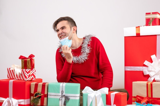 Front view young man taking off his mask sitting around xmas gifts