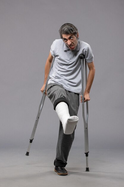 Front view young man using crutches due to broken foot on grey desk disable broke broken accident leg foot