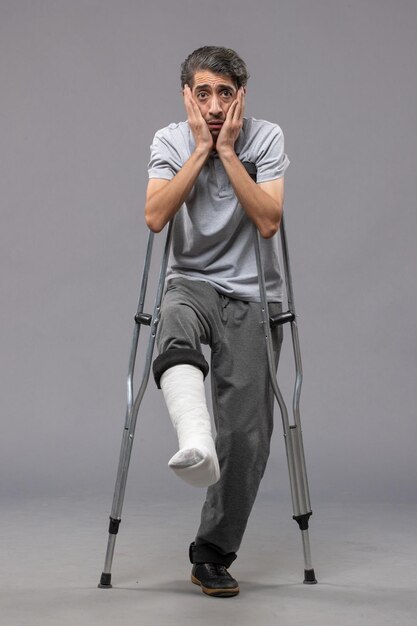 Front view young man using crutches due to broken foot on grey desk disable broke leg broken accident